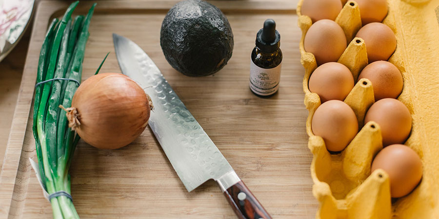 a kitchen knife, a spring onion, a white onion, an avocado, a small bottle of CBD oil and several eggs on top of a wooden chopping board