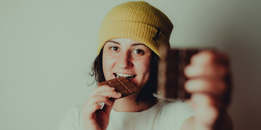 woman wearing a yellow beanie and white t-shirt biting a chocolate between her teeth and holding another bar of chocolate with her other hand